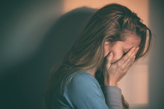 side profile of womans face, hiding in her hands, head slightly tilted forward. she has light brown long hair, wearing a long sleeves blue tshirt. background behind her is shadowed. 