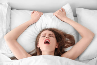 Woman lying in bed with eyes closed, gripping a pillow and appearing to be in a moment of intense pleasure or release.