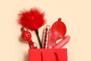 Red coloured sex toys including vibrator, gag ball, feather tickler and face mask. All placed inside a red gift bag on peach coloured background.