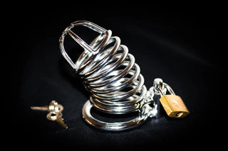 Close-up of a chrome male chastity cage with a padlock and keys, placed on a black background, symbolising control and restraint in BDSM play