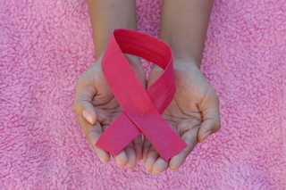 Hands holding a pink ribbon on a pink textured background, symbolizing breast cancer awareness.