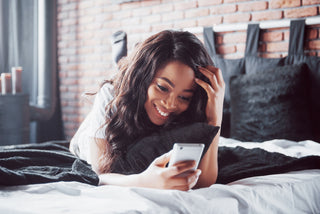 woman laying on her belly on a bed with her feet up behind her. she is looking into her phone, smiling. she has brown long curly hair.
