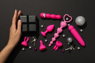 an image of pink sex toys placed flat lay on black background. on the left side is a left female hand, sideways, holding a small black gift box. 