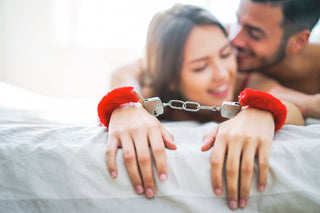Smiling woman lying on a bed wearing red fluffy handcuffs, with a man leaning close behind her in an intimate, playful moment.