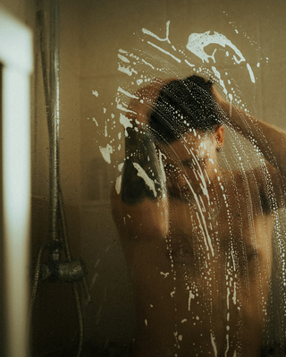 Person showering behind a glass door covered in water droplets and soap suds, with light filtering into the bathroom