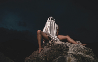 Person wearing a ghost costume made of shiny fabric with sunglasses, sitting on rocks at night with legs exposed.