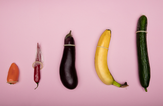Row of vegetables and fruits with condoms on them against a pink background, symbolizing safe sex and size variety.