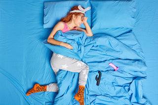 Woman sleeping contently in a bed with bright blue sheets, wearing fun pajamas and leopard-print socks, with two vibrators lying beside her.