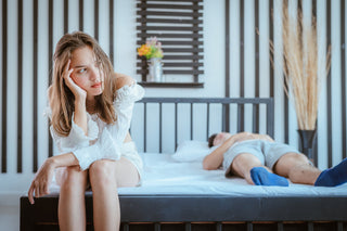Woman sitting at the edge of a bed looking bored or frustrated while a man sleeps behind her, suggesting post-sex dissatisfaction or emotional disconnect.
